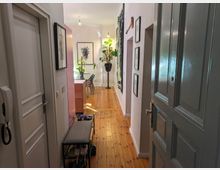 Blick in einen hellen Flur mit Holzboden und weißen Wänden, dekoriert mit Pflanzen, Bildern und einem an der Wand hängenden Teppich. Im Vordergrund befinden sich eine geöffnete graue Tür, ein Schuhregal mit Sitzbank und Wandbilder; am Ende des Flurs steht ein Tisch mit Stühlen.