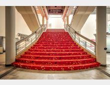 Breite Treppe mit rotem Teppich, verziert mit floralen und geometrischen Mustern, in einem offenen Innenbereich eines Kinos. Auf beiden Seiten befinden sich Metallgeländer, und oben ist ein Schild mit der Aufschrift „Zu den Kinos 6–10“ sichtbar.