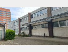 Innenhof vor einem flachen, zweigeschossigen Büro- oder Industriegebäude mit grauer Putzfassade, langen Fensterbändern und einem auskragenden Vordach über dem Eingang. Vor dem Gebäude stehen Metall-Fahrradbügel auf einer gepflasterten Fläche, daneben Büsche und Kletterpflanzen; im Hintergrund ragt ein hoher Backsteinbau auf.