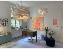 Heller Raum mit moderner und kreativer Einrichtung, darunter ein Holzschreibtisch, große organisch geformte Hängelampen, sowie bunte Wandkunst und Pflanzen. Ein Fenster mit Dekorationen und ein helles Sideboard ergänzen das Ambiente.