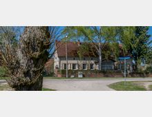 Ein zweigeschossiges Wohnhaus mit rotem Ziegeldach und beige Fassade, umgeben von Bäumen und Hecken. Im Vordergrund ein Baumstamm und ein Straßenschild mit den Namen "Gebhardstraße" und "Seegerallee".