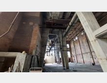 Innenansicht einer verlassenen Industriehalle mit hohen Backsteinwänden, Betonstützen und einem Fragment einer Metallkonstruktion an der Decke. Der Boden ist staubig und übersät mit Schutt, und Tageslicht dringt durch hohe Fenster ein.