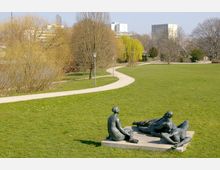 Weitläufiger Park mit einer grünen Rasenfläche, durchzogen von geschwungenen Spazierwegen und umgeben von kahlen und blatttragenden Bäumen. Im Vordergrund eine Skulpturengruppe aus Bronze auf einem Sockel, im Hintergrund sind mehrstöckige Wohngebäude zu erkennen.