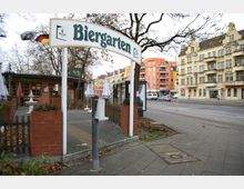 Ein Biergarten mit einem großen Eingangsschild aus Metall, umgeben von roten Ziegelmauern, kleinen Holzzäunen und Sonnenschirmen. Im Hintergrund sind mehrstöckige Wohnhäuser, eine Straße mit Autos und ein Wartehäuschen für den öffentlichen Nahverkehr zu sehen.