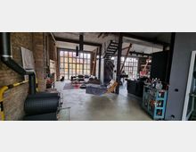 Industrieller Wohnraum mit hohen Decken, großzügigen Fenstern und freiliegenden Ziegelwänden; sichtbare Einrichtung umfasst eine Lounge-Ecke mit Sofas, eine Hängeliege, eine offene Metalltreppe und einen Holzofen. Im Hintergrund sind Holzbalken und ein Blick auf ein gelbes Gebäude durch die großen Fenster zu sehen.