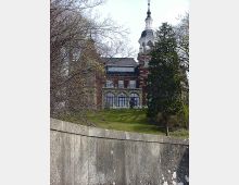Historisches Gebäude mit rotem Ziegelmauerwerk, weißen Fensterrahmen und einem markanten Turm mit Kuppel. Das Gebäude steht auf einer leicht erhöhten Grünfläche, umgeben von Bäumen, mit einer Betonmauer im Vordergrund.