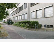 Straßenszene mit einem grauen, mehrstöckigen Gebäude mit geschlossenen Rollläden an den Fenstern. Vor dem Gebäude befinden sich ein Bürgersteig, eine Glas-Wartehäuschen und gepflegte Grünflächen mit niedrigen Büschen.