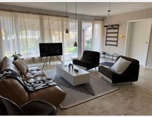 Wohnzimmer mit großen Fenstern, die Blick auf einen Garten bieten. Der Raum ist mit einem braunen Ledersofa, zwei schwarzen Sesseln, einem weißen Couchtisch und einem Fernseher auf einem Stativ möbliert. Helle Vorhänge und cremefarbene Fliesen bestimmen das Ambiente.