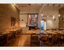 Gemütlicher Speiseraum mit Backstein- und weiß gestrichenen Wänden, Holzböden und warmem Licht. Große Holztische mit gedeckten Plätzen, Pendelleuchten an der Decke sowie ein Getränkekühlschrank bilden die Hauptmerkmale.