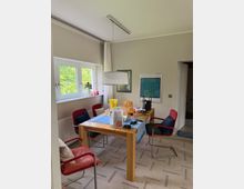 Esszimmer mit rechteckigem Holztisch, blauem Tischläufer und vier rot gepolsterten Stühlen; darüber hängt eine große, zylindrische Pendelleuchte. Links befindet sich ein Doppelfenster mit weißen Vorhängen und Blick ins Grüne, an der Wand hängen gerahmte Bilder und rechts führt eine offene Tür in einen angrenzenden Flur.