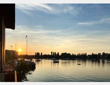 Sonnenuntergang an einem Fluss mit ruhigem Wasser, auf dem mehrere Boote treiben. Im Vordergrund ist ein Segelboot und der Balkon eines Gebäudes sichtbar, während am Horizont Bäume, ein Wasserturm und ein Kran zu erkennen sind.