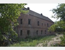 Backsteinbau eines verlassenen Gebäudes mit teilweise zerstörten oder fehlenden Fenstern, umgeben von spärlicher Vegetation und Bäumen. Das Haus hat ein Satteldach und befindet sich in einer verwilderten Umgebung.