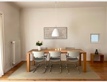 Esszimmer mit einem rechteckigen Holztisch, umgeben von sechs Stühlen mit Metallgestell und hellen Sitzflächen. Über dem Tisch hängt eine weiße Hängeleuchte, an der Wand dahinter ein abstraktes Kunstwerk; links ein Heizkörper, rechts ein kleines Fenster und ein Holztisch mit einer Vase.