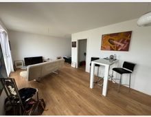 Wohnzimmer einer modernen Wohnung mit hellem Holzboden, einem beigen Sofa vor einem Fernseher, einem hohen weißen Esstisch mit zwei schwarzen Barhockern und abstrakter Kunst an der Wand. Im Raum befinden sich außerdem einige Bilderrahmen und Alltagsgegenstände.