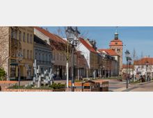 Fußgängerzone mit historischen Gebäuden und roten Ziegeldächern, rechts ein markanter Kirchturm aus Stein. Im Vordergrund befinden sich Bänke, Laternen und eine moderne Metallskulptur, umgeben von jungen Bäumen und bepflanzten Beeten.