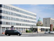 Ein modernes Verwaltungsgebäude mit einer schlichten, weißen Fassade und zahlreichen Fenstern; im Hintergrund ist die grün-goldene Kuppel einer Kirche oder eines Doms sichtbar. Im Vordergrund verläuft eine Straße mit einem schwarzen Van und weiteren Fahrzeugen.