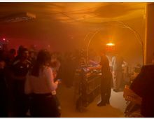 Eine dunkle Indoor-Party- oder Konzertszene mit orangem Licht, auf der Personen vor einer DJ- oder Musikstation tanzen. Im Hintergrund steht ein DJ-Pult mit Geräten, und eine Hängelampe sorgt für gedämpfte Beleuchtung.