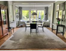 Esszimmer in einem stilvollen Wohnbereich mit einem runden Esstisch und vier Stühlen auf einem großen Teppich. Im Hintergrund ein heller Sitzbereich mit bodentiefen Fenstern und Blick in einen Garten; Wände sind dezent dekoriert mit Bildern und Spiegeln.