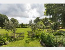 Ein ländlicher Garten mit einem kleinen Holzzaun, der eine grüne Rasenfläche und Gemüsebeete umgibt. Im Hintergrund sind Bäume und Büsche zu sehen, während der Himmel bewölkt ist.