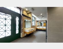 Moderner Waschraum mit Backsteinwänden, großen Fenstern mit grünem Rahmen und mehreren rechteckigen Waschtischen mit Spiegeln. Der Boden ist mit dunklen Fliesen ausgelegt, und das natürliche Licht fällt durch die Fenster ein.