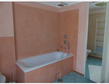 Badezimmer mit einer Badewanne in einer Nische, umgeben von orangefarbenen Wänden. Auf dem Boden sind grüne Fliesen, und im Hintergrund ist ein Handtuchheizkörper zu sehen.