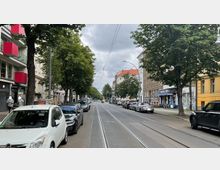Straßenansicht einer urbanen Umgebung mit parkenden Autos entlang beider Seiten der Straße, Straßenbahnschienen in der Mitte und Bäumen, die den Gehweg säumen. Links befinden sich moderne Gebäude mit roten Balkonen, rechts ältere Fassaden sowie Geschäfte und Cafés.