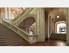 Großzügiger historischer Treppenbereich mit verzierten Balustraden, hohen Arkaden und einer geschwungenen Decke. Im Hintergrund befindet sich ein Eingangsbereich mit Glastüren und modernem Lichteinlass.