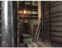 Industrieanlage mit großen metallischen Zylindern und sichtbaren Rohren an der Decke. Im Hintergrund ist eine Tür mit einem Warnschild („Vorsicht!“) sowie eine Metallleiter zu sehen.