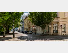 Pflastersteinstraße in einer Stadt mit historischen Gebäudezugen, darunter ein gelbliches Gebäude mit Ornamentfassade. Links stehen Bäume mit Schatten, rechts parken Autos, und ein Straßenschild weist auf eine Parkzone hin.