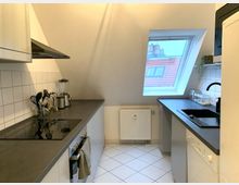 Moderne Einbauküche in einer Dachgeschosswohnung mit einer Dachschräge. Zu sehen sind eine Arbeitsplatte mit Cerankochfeld, ein Spülbecken, weiße Oberschränke, eine schräge Fensteröffnung mit Blick auf Dächer, sowie weiße Bodenfliesen und Wandfliesen in der Küchenzeile.