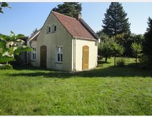 Kleines, freistehendes Häuschen mit hellem Putz, rotem Ziegeldach und zwei Holzläden an den Türen, neben einem Anbau. Es steht in einem grünen Garten mit Rasen, Zaun und hohen Bäumen im Hintergrund unter blauem Himmel.