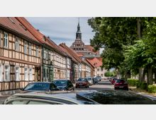 Straßenszene in einer deutschen Altstadt mit Fachwerkhäusern auf der linken Seite, einer gepflasterten Straße, Autos am Straßenrand und grünen Bäumen auf der rechten Seite. Im Hintergrund ragt ein Kirchturm mit markanter Architektur hervor.