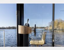 Steuerstand eines Bootes mit modernem Lenkrad und Schalthebel, umgeben von großen Fenstern mit Blick auf einen See und Anlegestellen. Im Hintergrund sind Bäume und ein klarer Himmel zu sehen.
