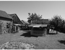 Schwarz-Weiß-Aufnahme eines ländlichen Gartens mit Wiese, einem kleinen Schuppen mit Wellblechdach, Obstbäumen und einem Ziegelgebäude links im Hintergrund. Im Vordergrund ist ein Gemüsebeet zu sehen, umgeben von einer ruhigen, offenen Umgebung.