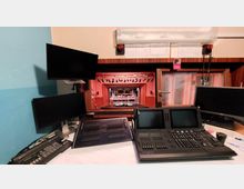 Blick aus einem Licht- und Tontechnikraum auf eine Bühne mit Scheinwerfern und Bühnenelementen. Im Vordergrund befinden sich Bedientafeln, Monitore und Steuerpulte für die Technik, daneben ein Fenster mit Schalldämmung.