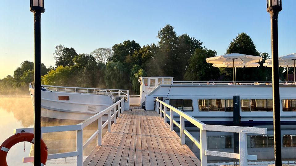 Holzsteg mit Geländer, der zu einem festgemachten Schiff führt, flankiert von zwei Laternen. Im Hintergrund Wasser, Bäume und ein klarer, blauer Himmel.