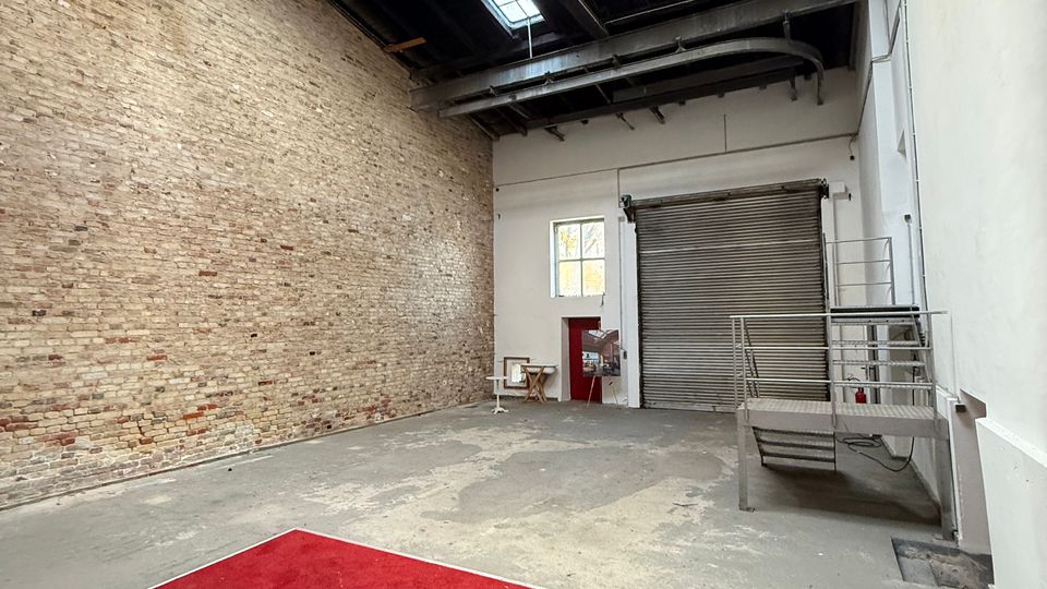 Leere Industriehalle mit hoher Decke, sichtbaren Stahlträgern und Oberlichtern; links eine freiliegende Ziegelwand und rechts weiße Wände. Am Ende befinden sich ein großes Rolltor, ein kleines Fenster und eine Metallplattform mit Treppe; im Vordergrund liegt ein roter Teppich auf dem Betonboden.