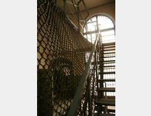Metalltreppe in einem Gebäude mit hohen, vergitterten Fenstern; an der Seite der Treppe ist ein Sicherheitsnetz aus Metall angebracht. Im Hintergrund sind Wände und ein Türdurchgang zu sehen.
