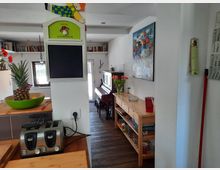 Wohnraum mit offener Küche und Holzfußboden. Im Vordergrund stehen eine Küchenzeile mit Toaster und Obstschale sowie eine Kreidetafel. Im Hintergrund sind ein Klavier, ein Sideboard mit Dekoration und Bücherregale an der Wand zu sehen.