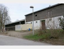 Außenansicht eines Gebäudekomplexes mit gemischter Fassade aus Holz und weißem Putz, ergänzt durch einen kleinen gelben Container. Ein gepflasterter Weg, eine Laterne, blattlose Sträucher und ein kahler Baum sind im Vordergrund erkennbar.