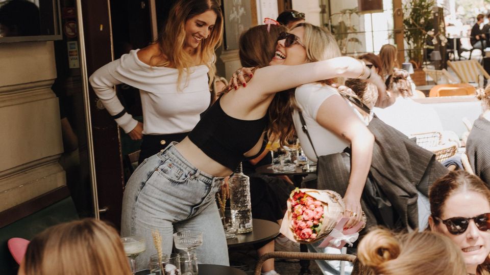 Außenbereich eines Straßencafés mit dicht stehenden runden Tischen und geflochtenen Stühlen unter Markisen. Über dem Eingang hängt ein schwarzes Schild mit der Aufschrift „BAR 1000 GRAHAM“, daneben sind Pflanzen und eine belebte Gehwegterrasse zu sehen.