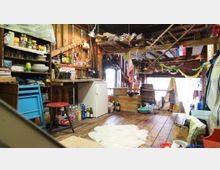 Rustikaler Innenraum einer Holzhütte mit einer offenen, improvisierten Küchenecke. Regale mit Geschirr und Lebensmitteln, ein kleiner Kühlschrank sowie hölzerne Dekorationen und Haushaltsgegenstände dominieren den Raum; im Hintergrund sind eine geöffnete Tür und Fenster, die Tageslicht hereinlassen.