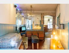 Gemütliche Café-Innenansicht mit Vintage-Möbeln, darunter eine grüne Couch, Holzbänke und ein rustikaler Kaffeetisch. Eine alte Röhre-TV steht in einer Ecke, umgeben von warmen Lichtquellen und Wänden mit hölzernem Wandpaneel, ergänzt durch einen offenen Durchgang mit sichtbarer Steinmauer.