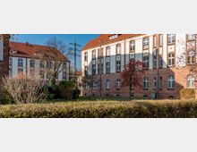 Backsteingebäude mit weißen Fassadenelementen und großen Fenstern, umgeben von einem gepflegten Garten mit Bäumen und Büschen; im Hintergrund ein klarer Himmel und ein Strommast.