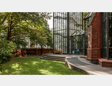 Moderne Glasfassade eines Gebäudes mit einer roten Backsteinstruktur daneben, umgeben von einer Grünfläche mit Bäumen und einem kleinen gepflasterten Bereich mit Sitzbank. Im Hintergrund sind weitere Gebäude und eine niedrige Ziegelmauer zu sehen.