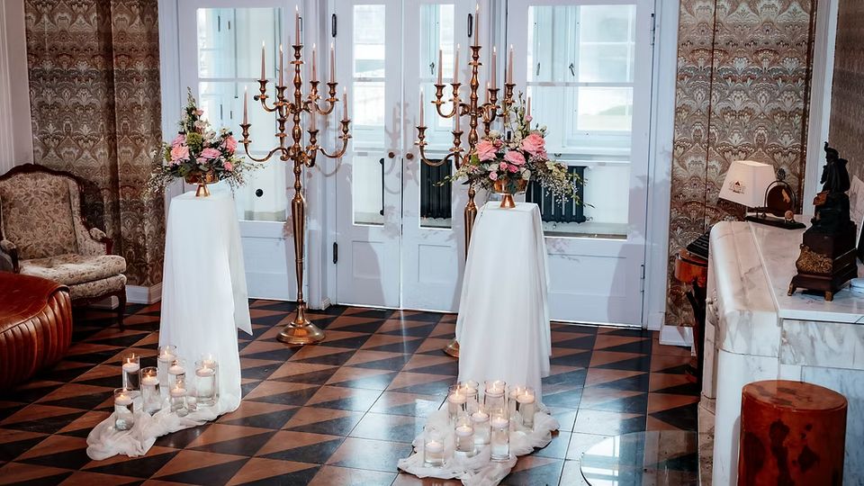 Eleganter Innenraum mit weiß gerahmten Glastüren, gemusterter Tapete und schwarz-braun kariertem Fliesenboden. Im Vordergrund stehen zwei mit weißen Tüchern bedeckte Stehtische mit Blumenarrangements, flankiert von goldenen Kerzenständern und Gruppen kleiner Kerzen in Glasgefäßen.