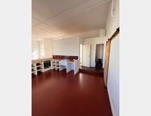 Küche in einem offenen Raum mit roten Bodenfliesen, weiß gestrichenen Wänden und heller Decke. Die Küche verfügt über weiße Schränke mit offenen Regalen, eine Arbeitsfläche, rote Fliesen als Rückwand, einen eingebauten Ofen und Wasserhahn sowie eine angrenzende Tür zu einem anderen Bereich.