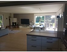 Offener Wohn-Essbereich in einem modernen Haus mit heller Fliesenboden, einem großen Fernseher an der Wand, einem Kamin in der Ecke und großen Glas-Schiebetüren mit Blick auf eine Terrasse und einen Garten. Rechts im Vordergrund ist eine Küchenarbeitsplatte mit Schränken zu sehen.