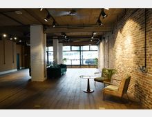 Ein industriell gestalteter Innenraum mit einer Mischung aus Backstein- und weiß gefliesten Wänden, freiliegender Decke mit Beleuchtung und Holzboden. Der Raum ist mit modernen grünen Polstermöbeln, einem kleinen Tisch und Pflanzen dekoriert und verfügt über große Fenster, die natürliches Licht hereinlassen.