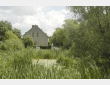 Ein graues Wohnhaus mit Satteldach steht in einer grünen, ländlichen Umgebung, umgeben von hohen Gräsern und Bäumen; im Vordergrund befindet sich ein kleiner Teich mit Wasserpflanzen. Im Hintergrund führt ein Weg von der Vegetation gesäumt weiter.